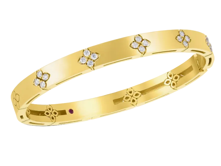 A yellow gold bangle bracelet with a continuous floral pattern, featuring small clear pavé diamonds in four-flower motifs and a tiny red gemstone accent on the inner band, shown on a light gray background.