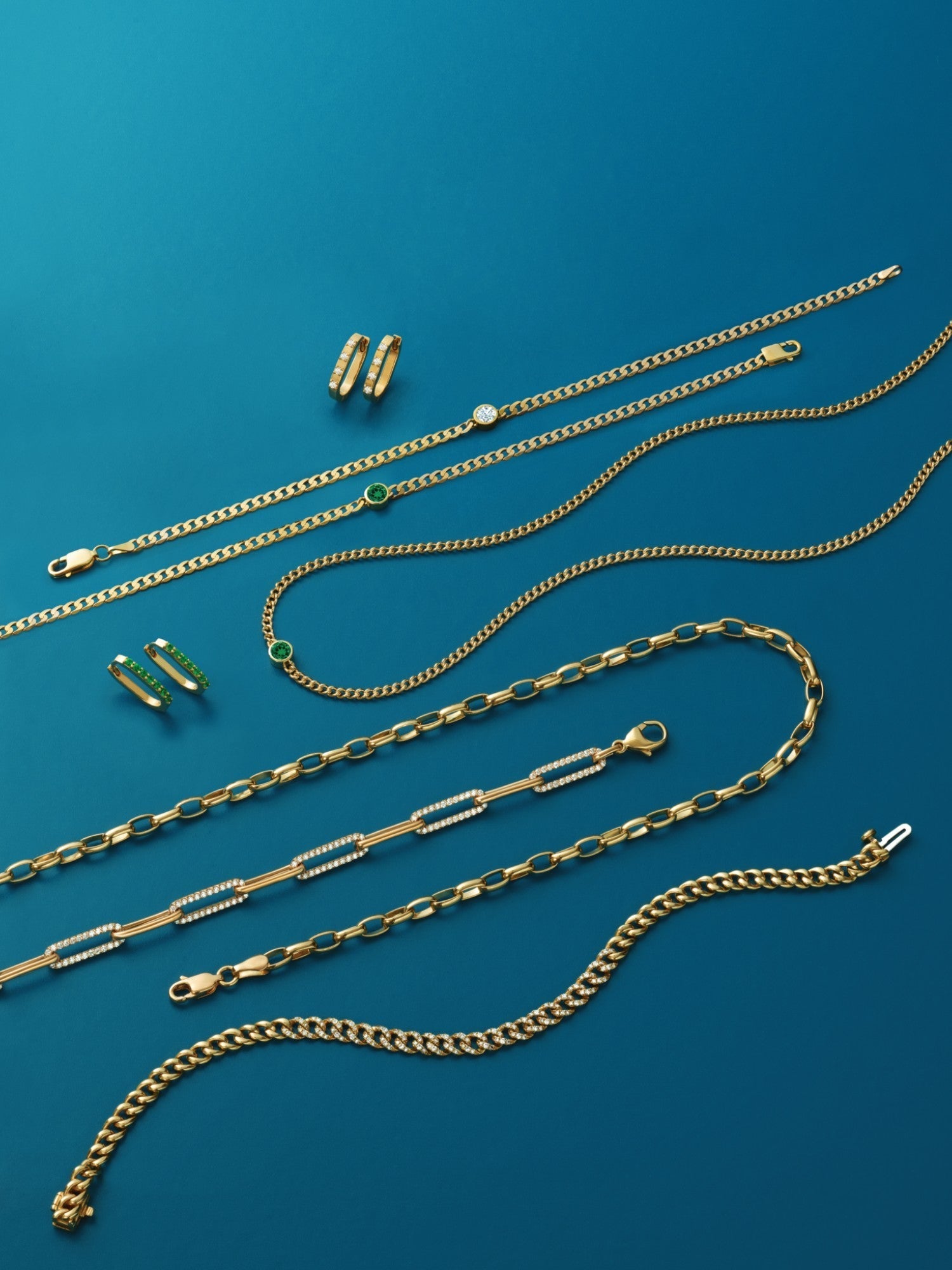 **Alt Text:** A collection of elegant gold necklaces, chains, and huggie hoop earrings displayed on a deep blue background. Some pieces feature diamond and emerald accents, with intricate chain designs and polished gold finishes.