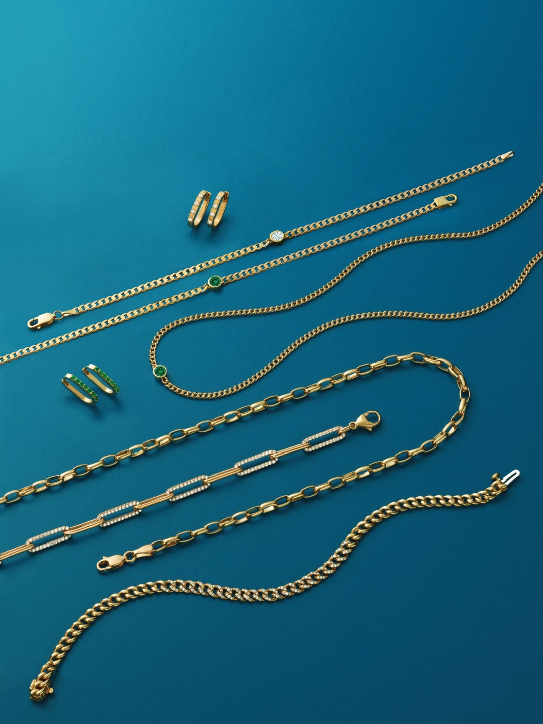 **Alt Text:** A collection of elegant gold necklaces, chains, and huggie hoop earrings displayed on a deep blue background. Some pieces feature diamond and emerald accents, with intricate chain designs and polished gold finishes.