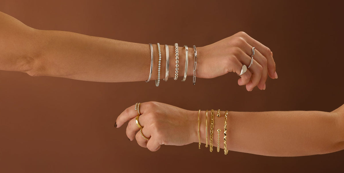 Two human arms and hands shown against a warm brown studio background, each wearing multiple thin bracelets and rings. One wrist has silver-toned bracelets, and the other has gold-toned bracelets and rings.